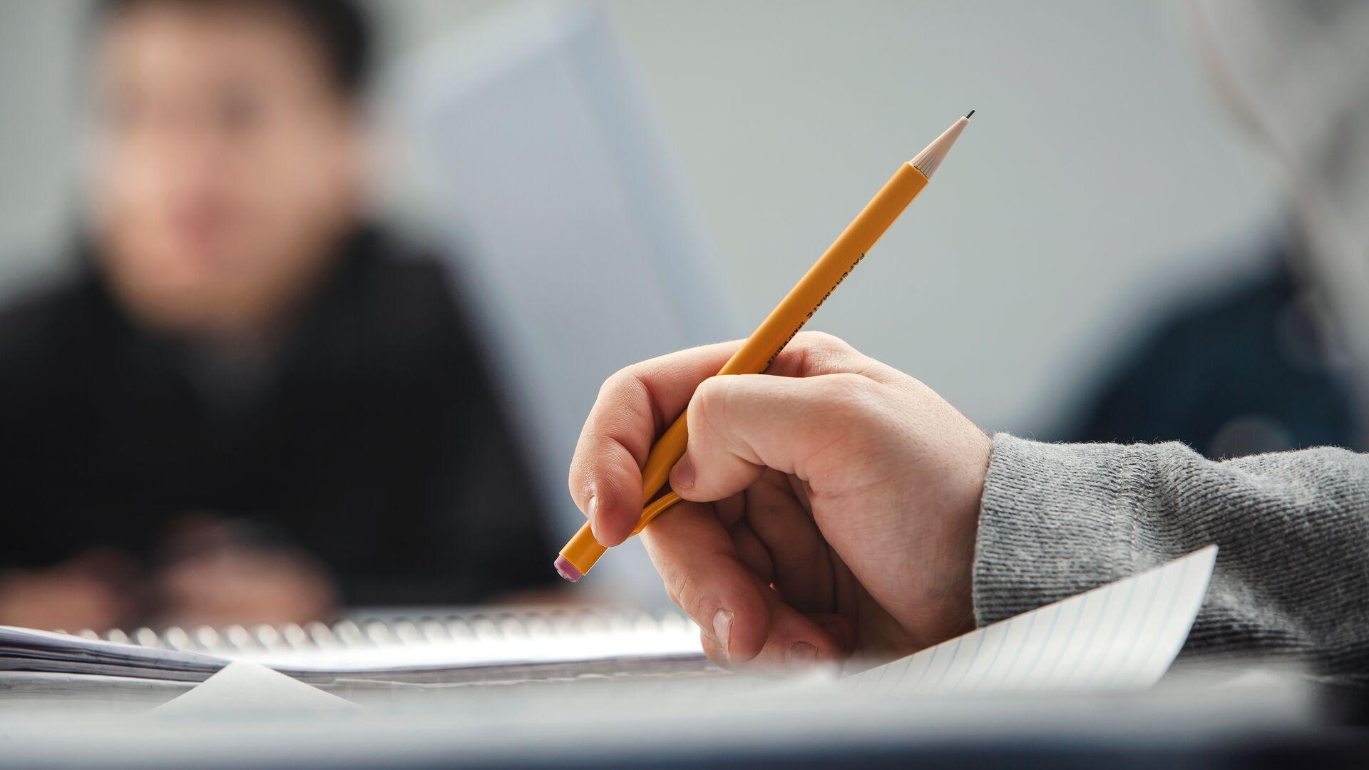 Hand holding a pencil over a notebook, out of focus student in the background.